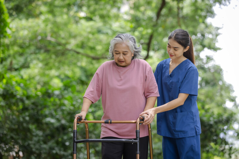 Elderly woman with walker carefully assisted by young nurse in a lush green park setting